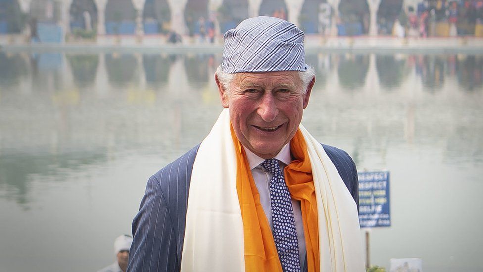 King Charles smiles at the camera as he wears a blue and white turban to cover his head and an orange and cream shawl around his shoulders on a visit to India.
