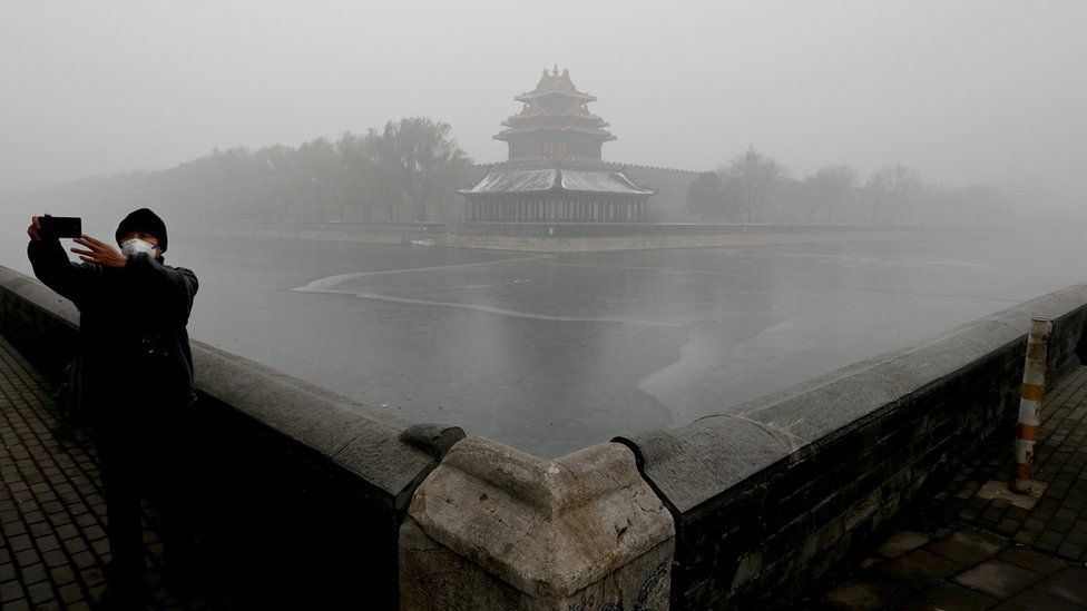 At the Forbidden City a tourist takes a selfie, while wearing a mask and surrounded in smog