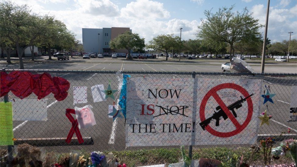 A sign at floral tribute at Parkland school reads "Now is the time" with a symbol of a gun crossed out