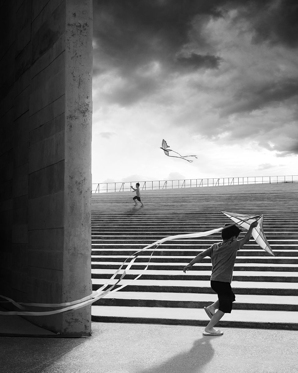 A black and white photo showing two children playing with kites on the steps of Teopanzolco Cultural Center in Mexico with dark clouds in the sky