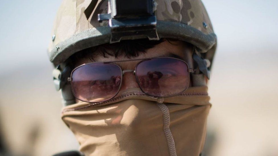 A members of the Triples pictured front on with his head in shot. He is wearing a combat helmet, sunglasses, and his mouth is covered