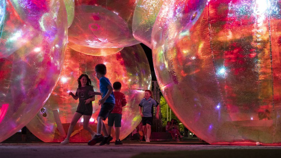 Children play among huge art installations resembling iridescent bubbles at dark