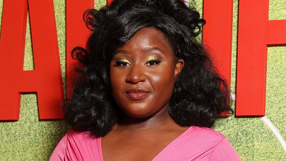 Susan Wokoma. Susan is a black woman in her 20s and is pictured at a red carpet event. Susan has shoulder-length curly black hair which is styled half-up-half-down. She has brown eyes and wears a gold eyeshadow and eye-liner make-up look. She wears a low-cut bubble gum pink dress and smiles at the camera