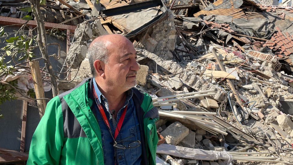 A man in green stands in front of rubble