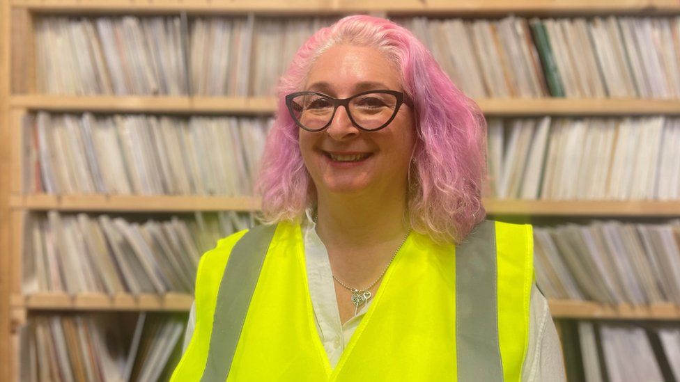 Karen Emanuel, CEO of Key Production. Karen is a white woman in her 50s with shoulder length curly hair which is dyed pink. She wears purple rimmed glasses and smiles at the camera. She's pictured on the factory floor in front of shelves of records. She's wearing a yellow high-vis vest over a white shirt.