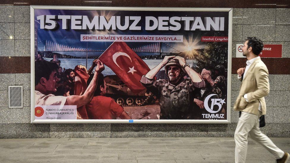 man in suit walking past billboard with image of soldier, Turkish flag and words 15 Temmuz Destani
