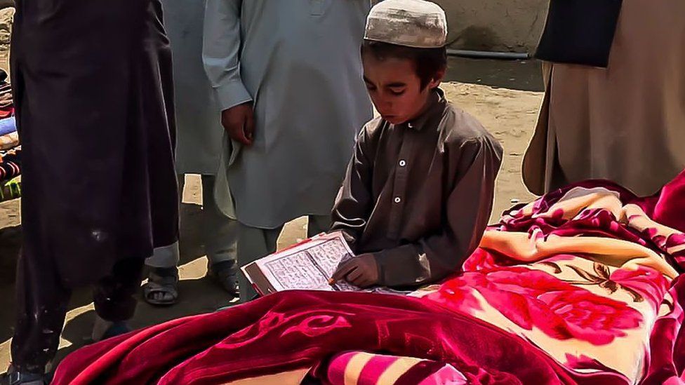 เด็กชายชาวอัฟกันอ่านหนังสือเกี่ยวกับศาสนาข้างๆ ศพของเหยื่อแผ่นดินไหวที่ถูกห่อด้วยผ้าห่มก่อนพิธีฝัง ซึ่งเสียชีวิตจากเหตุแผ่นดินไหวที่อำเภอกายัน จังหวัดปักติกา เมื่อวันที่ 22 มิถุนายน พ.ศ. 2565