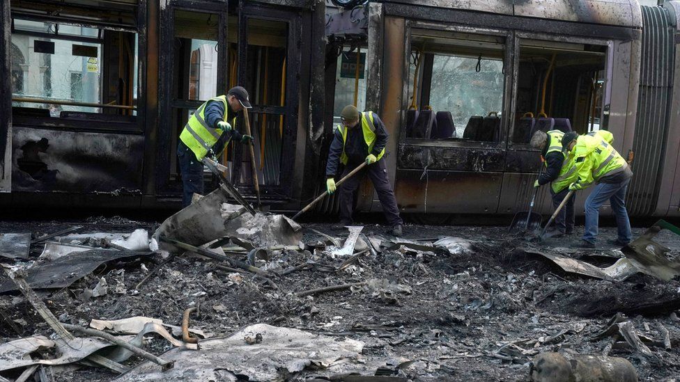 Vier Gemeindearbeiter räumen die verbrannten Trümmer einer Straßenbahn auf, die bei Unruhen im Stadtzentrum von Dublin in Brand gesteckt wurde
