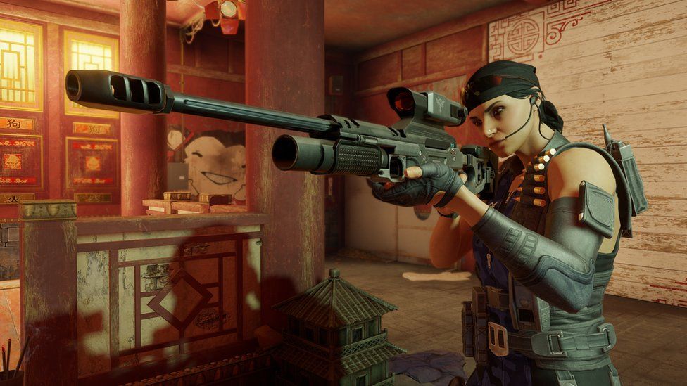 A female computer game character holds a sniper rifle up to her face - she's aiming the long barrel of the black weapon at an unseen foe, her eye looking down the red telescopic sight. She's wearing a bandana, communication earpiece and a tactical vest with spare bullet cartridges and other accessories attached to it. She's standing a room decorated in a red-hued, traditional Chinese style complete with wooden colums and intricate gold patterns on the walls.
