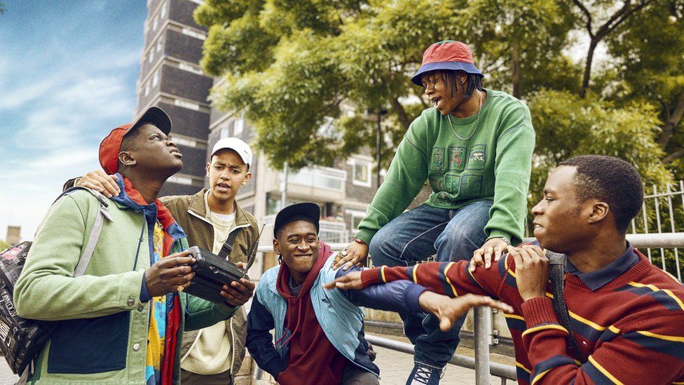 From left to right, the characters of Dane played by Yus Jamal Crookes - who is looking up - wearing a red cap and green jacket, holding a black radio, Junior played by Gabriel Robinson - who is wearing a brown jacket and white cap with his arm around Dane's shoulder, Bishop played by Tienne Simon - who is wearing a blue jacket with dark blue sleeves and a maroon hoody underneath with a dark coloured cap, reaching out his left arm, Kai played by Shanu Hazzan - wearing a green top and blue jeans with a silver chain around his neck and pink and blue bucket hat looking down at Tienne, and Bayo played by Juwon Adedokun, wearing a maroon sweater with blue and yellow lines, pointing towards Tienne. The background is of a large tower block covered by green trees.