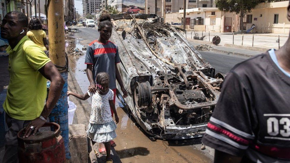 People walk past a burned out car in Dakar, on June 5, 2023, as protest calmed down four days after a court in Senegal sentenced opposition leader Ousmane Sonko, a candidate in the 2024 presidential election, to two years in prison on charges of 'corrupting youth' but acquitted him of rape and issuing death threats