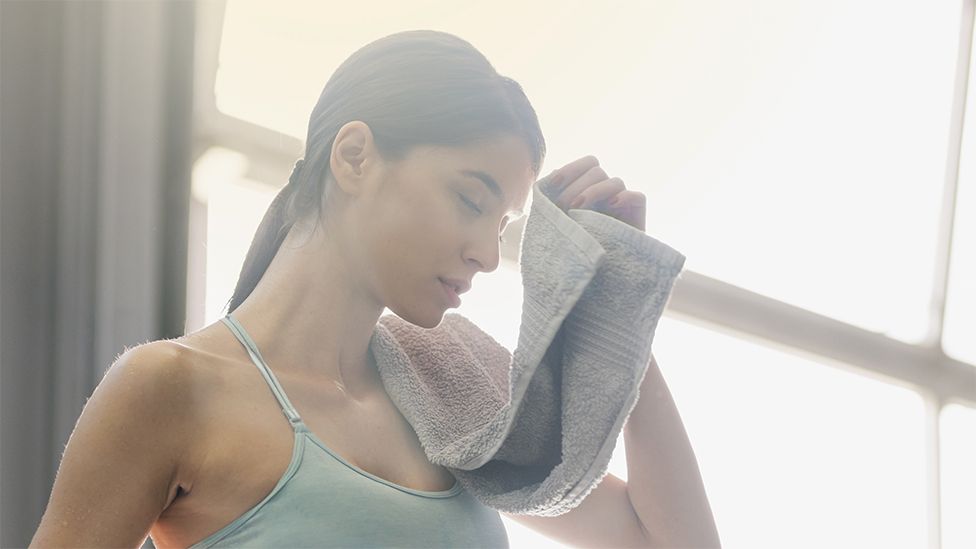 A woman wearing a light blue workout top, wiping sweat from her forehead after exercising. The towel she is using is grey, the background is at a window with the sun shining.