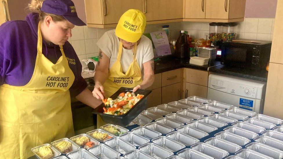 Becky in a purple t-shirt wearing a yellow apron and her mum Caroline put meal portions into containers.
