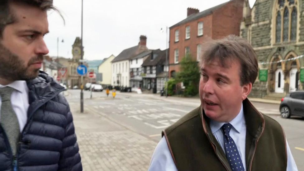 Craig Williams talks to BBC journalist while outside, wearing gillet, shirt and tie with a church and street in the background