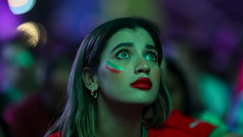 Женщина с зелено-красной краской на лице нервно смотрит на экран в футболке сборной Марокко