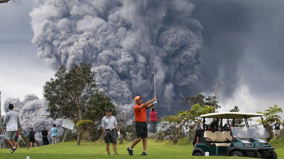 A man plays golf with a cloud from Kilauea behind him