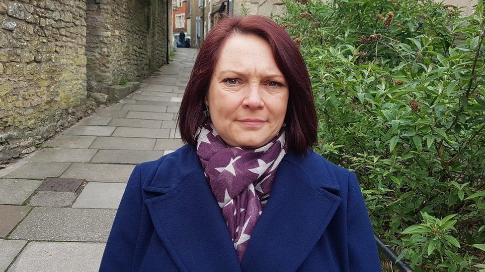 Kay wearing a blue coat and neck scarf standing in front of a bush and a stone building