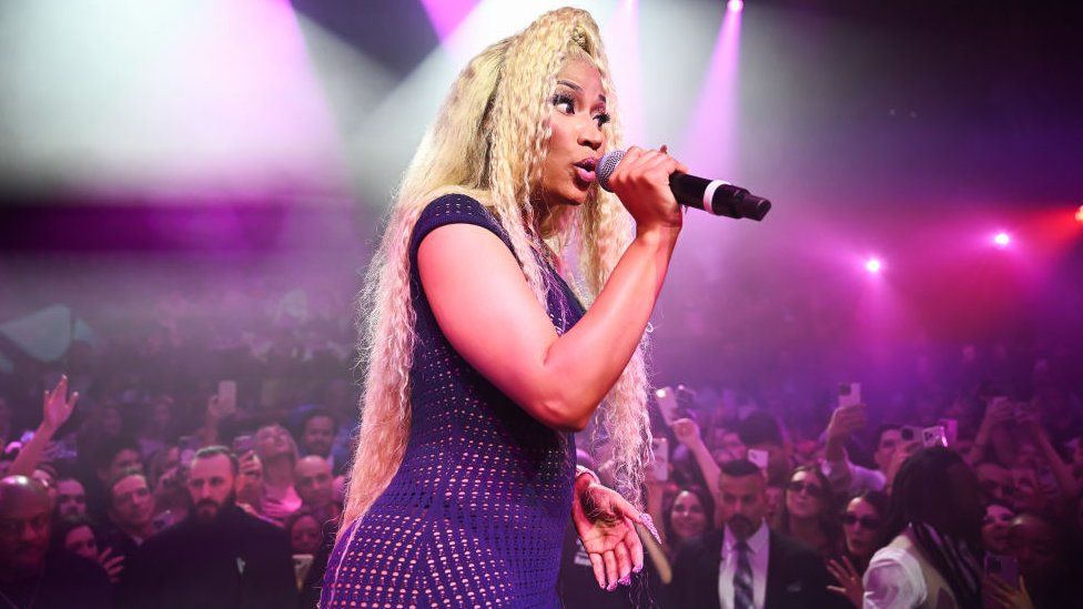Nicki Minaj performs on stage in December 2023. Nicki wears a large curly blonde wig and a stringy black dress. She is lit by pink lights and holds a microphone for her face. A crowd is visible behind her