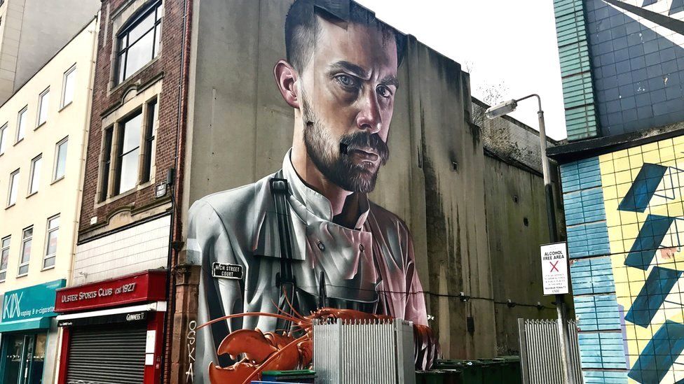 A large scale hyper-realistic portrait of a chef holding a lobster on a gable wall