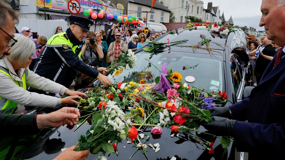 Люди возлагают цветы к катафалку, перевозящему гроб покойной ирландской певицы Шинед О'Коннор