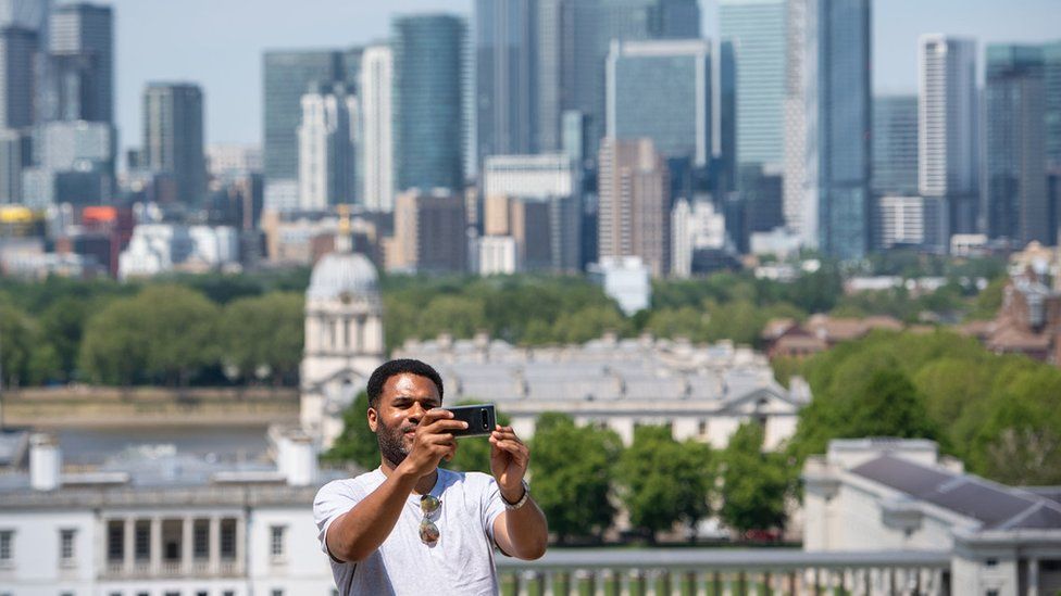 A man takes a selfie with the skyline of Canary Wharf, from the Royal Observatory in Greenwich Park, London