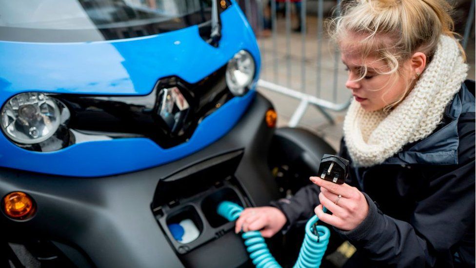 A woman holds a charging cable while crouching next to an electric vehicle
