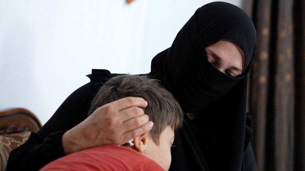 Alia is wearing a black niqab and is embracing her son. He is wearing a red t-shirt and has brown short hair.