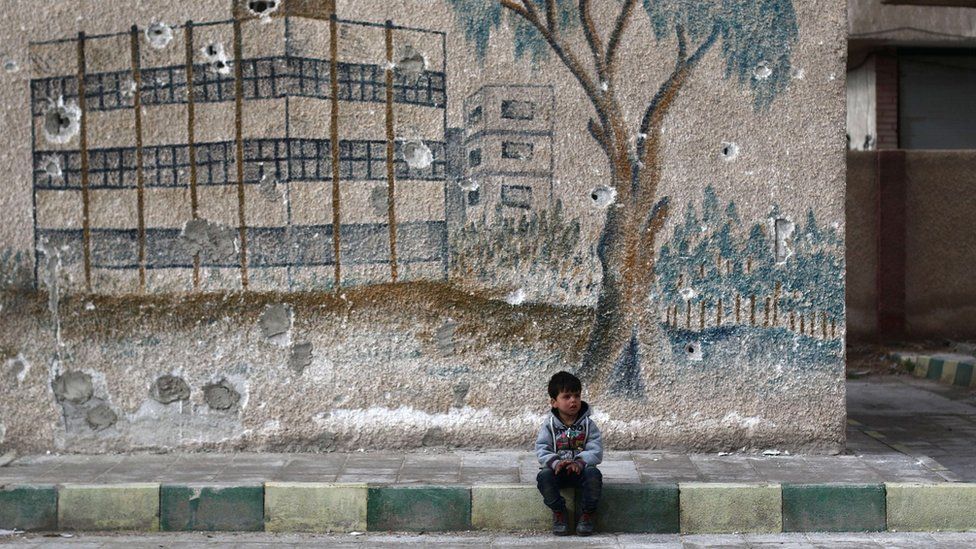A Syrian child sits in front of a mural covered in bullet holes on the wall of a former school in the rebel-held eastern Ghouta, on the outskirts of the Syrian capital Damascus, on January 5, 2016.