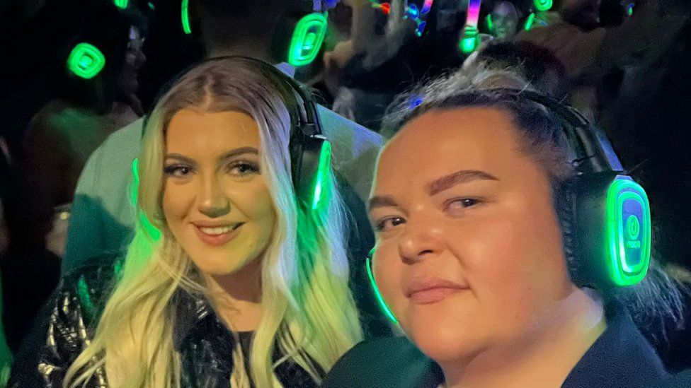 Nisha on the left and her friend, taking a selfie in which the two women are smling. The pair are wearing black headphones with green neon lights shining on its outer rims.