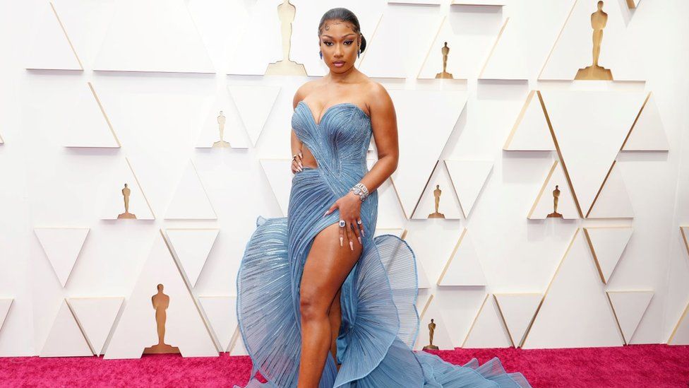 Megan Thee Stallion attends the 94th Annual Academy Awards at Hollywood and Highland on March 27, 2022 in Hollywood wearing one of Gaurav's designs. The dress has a pale blue bustier cascading down into a swirling train. It is embellished with silver gems and exposes Megan's side just above her hip as well as her left leg. The singer has one hand on her hip and has her black hair tied back in a low bun. Megan is pictured on the red carpet with a white and gold backdrop.