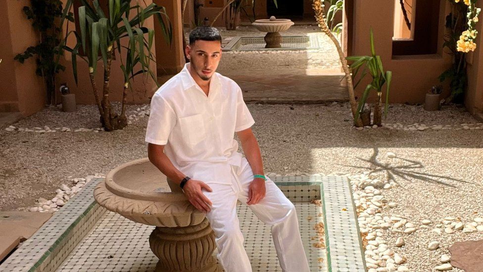 Islam Fessih sat on an ornate fountain in a courtyard posing for the camera. His hair is dark and styled quite short and he is a small dark moustache and beard. He is wearing a white short-sleeved shirt and white linen trousers and behind him in the courtyard there are stones on the floor and a few plants and another fountain in the background