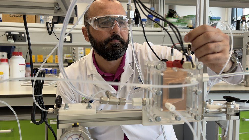 Prof Enrico Andreoli in a lab wearing a lab coat and safety glasses looking at an experiment
