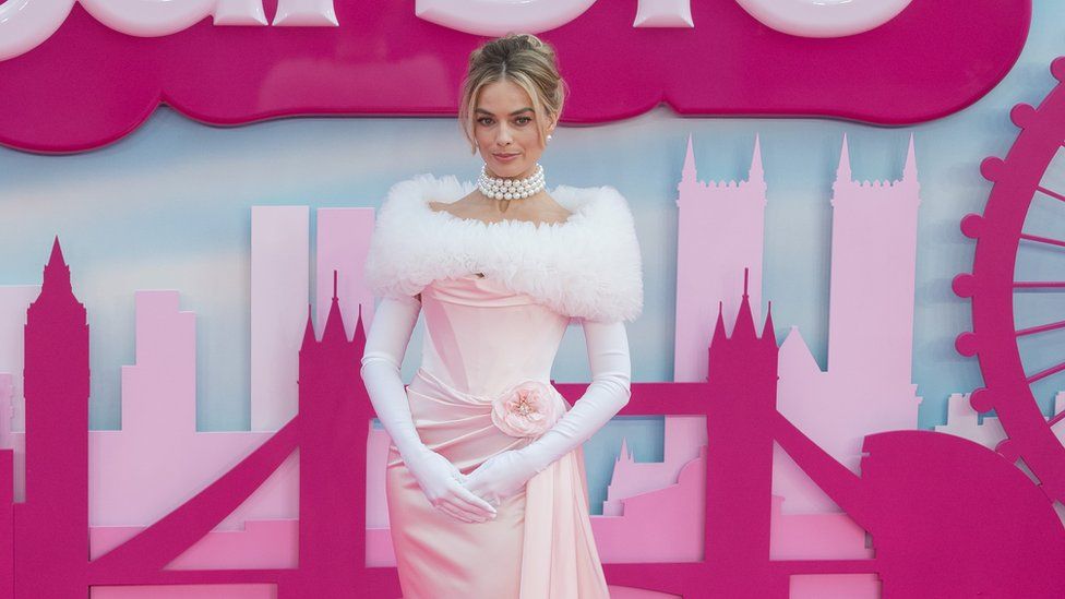 Margot Robbie at the London Barbie premiere, she is standing and smiling in front of a board which has a pink skyline of London, she is wearing a white fluffy shawl, pearl necklace and a light pink dress