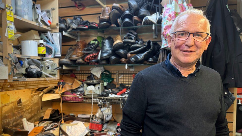 A man with glasses stands in his shoe repair shop, with shoes stacked behind him.
