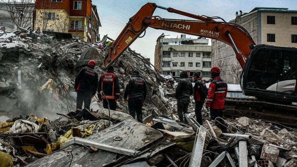 Workers watch as a digger sorts through the rubble after the earthquake struck in Malatya, Turkey