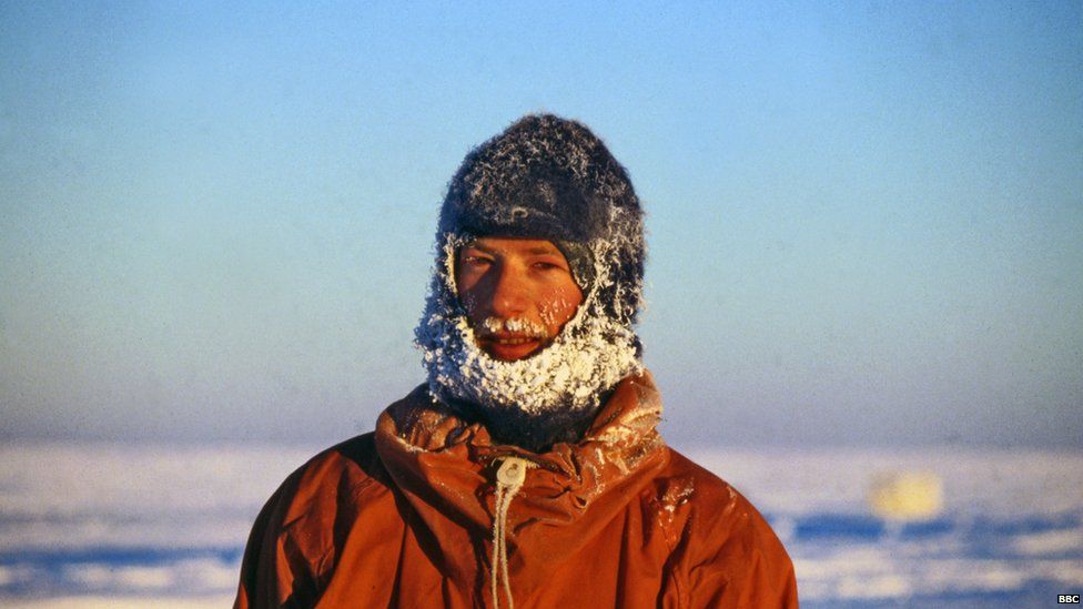 A young Peter Gibbs, wearing clothes covered in white snow. Blue sky behind.
