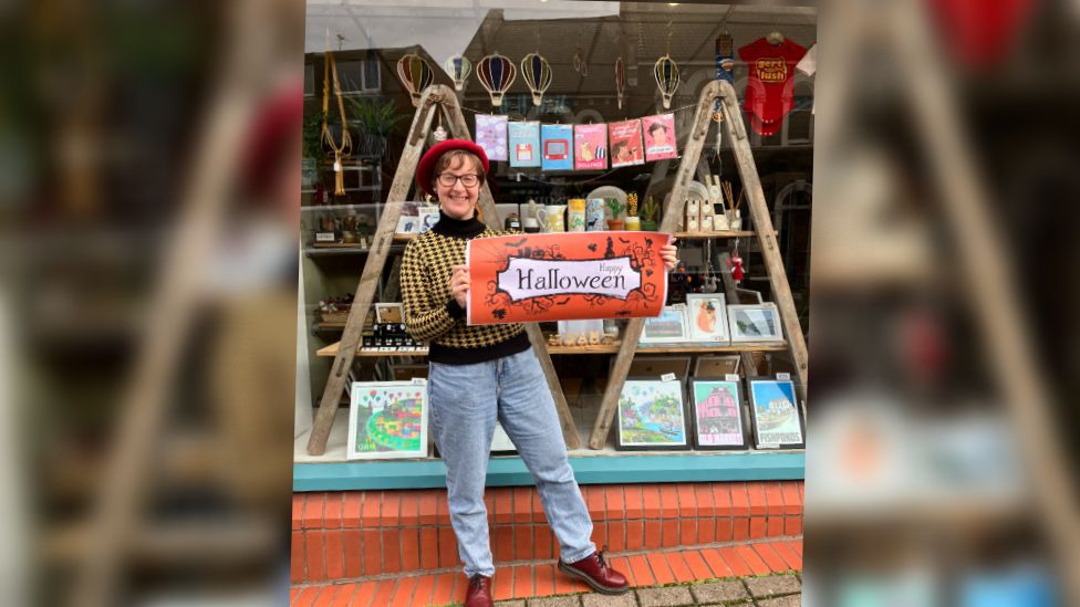 Nicola Bartlett outside her Eclectic Gift Shop holding an orange banner which advertises the Halloween trail.