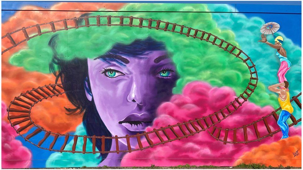 Street art by Ms Harris on Carfax street Swindon, it shows colourful clouds with a tightly bending train tack passing through the clouds. A large purple face with green eyes in the middle and Swindon's blondinie statue to the right hand side