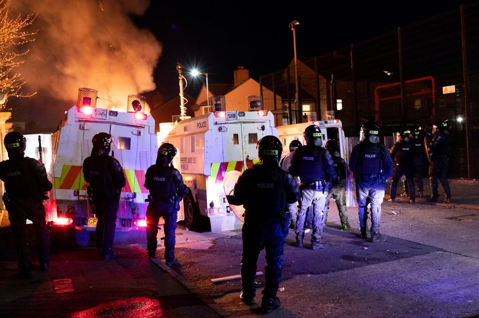 Police in riot gear watch as a car burns in front of police Land Rovers in Belfast