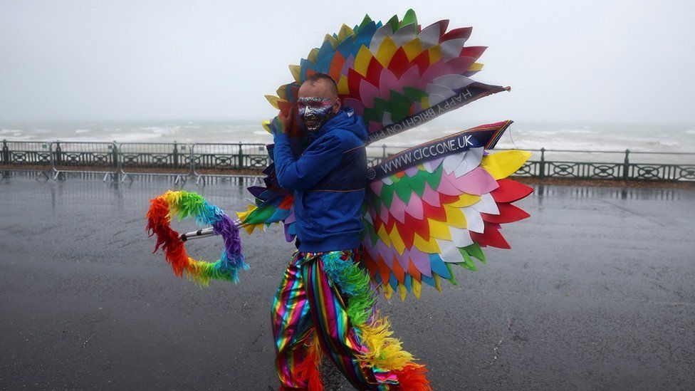 A man in a rainbow outfit battles the wind on Brighton seafront in front of choppy waves