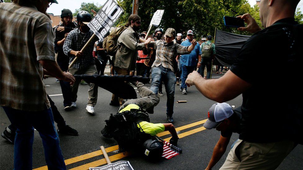a woman kicks a man who is on the ground clutching a US flag