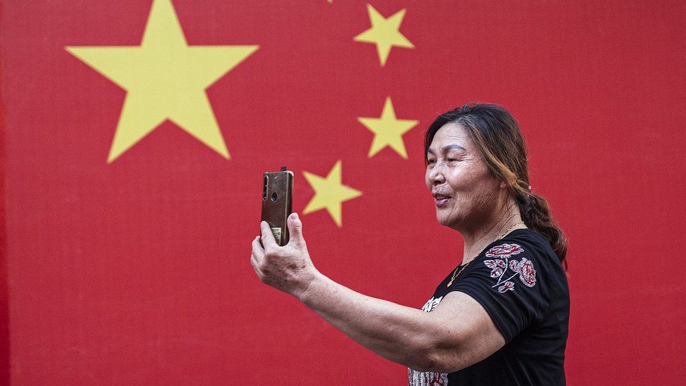 A women uses a iPhone to record a video near a wall of flags at EXPO garden park during the national holiday on 1 October, 2021 in Wuhan.