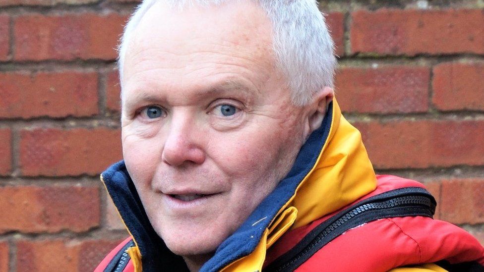 A man in a yellow RNLI jacket and red life preserver standing in front of a brick wall