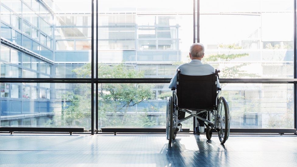 An old man in a wheelchair looking out of a window in an office corridor