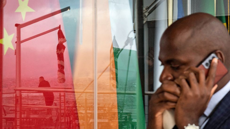 Мужчина разговаривает по мобильному телефону, проходя мимо отражения в окне другого мужчины, смотрящего на море, перед национальными флагами стран БРИКС (Китай, Индия, Россия, Южная Африка, Бразилия) страны в Кейптауне, ЮАР – июнь 2023 г.