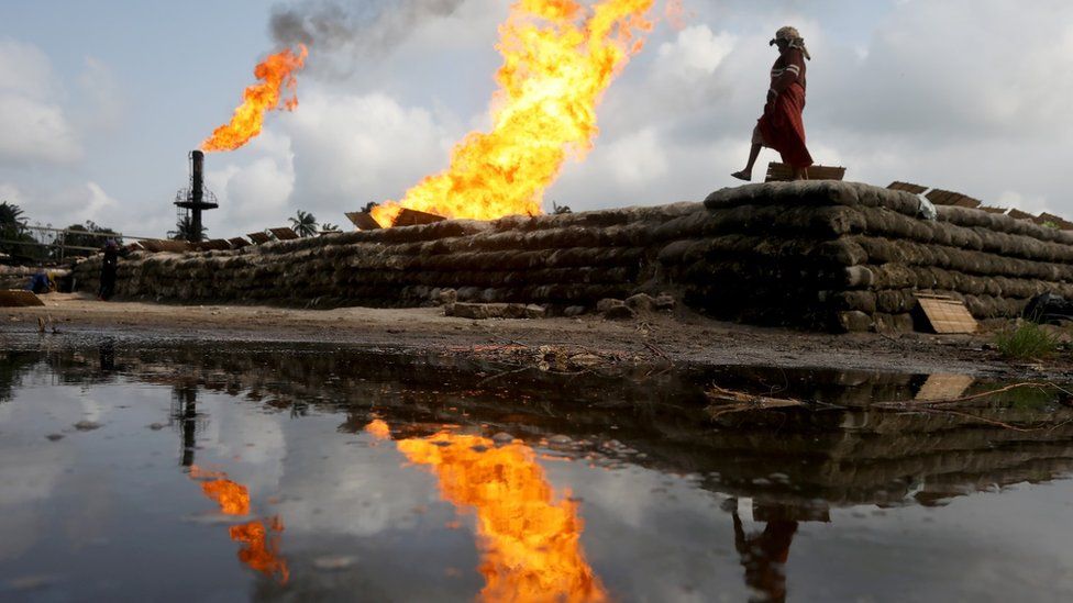 A reflection of two gas flaring furnaces and a woman walking on sand barriers is seen in the pool of oil-smeared water at a flow station in Ughelli, Delta State, Nigeria September 17, 2020.
