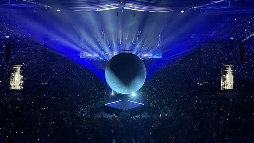 A photo showing Rosalie's blocked view - she is central in the crowd but quite far up in the stands and perfectly central is the inflatable moon which dominates the view of the stage. The prop is lit by blue lights and there are two screens either side of it showing The Weeknd performing.