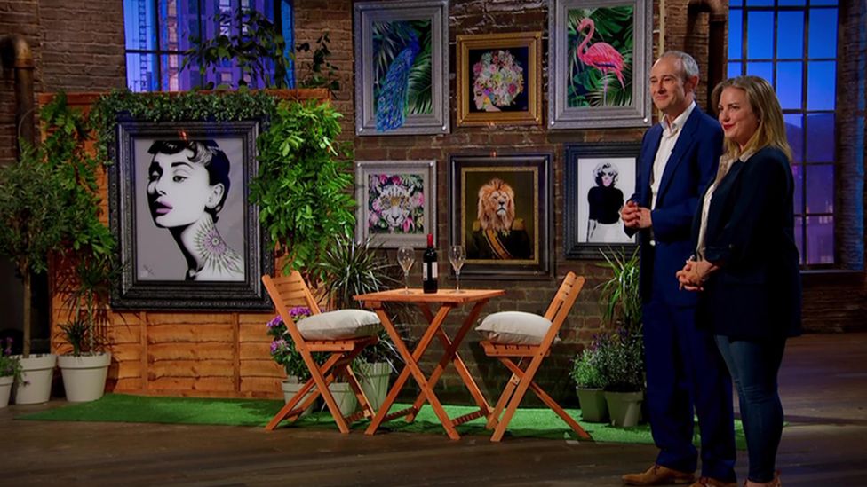Image of the Dragons' Den. A man and a woman standing in front of a table and chairs with artwork put up on the walls.