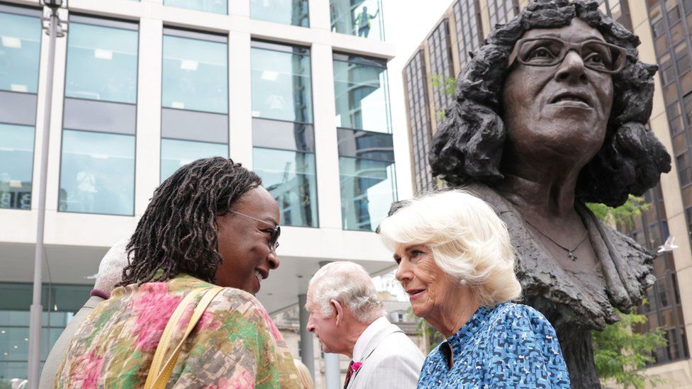 Camilla speaks to a woman in front of copper bust.