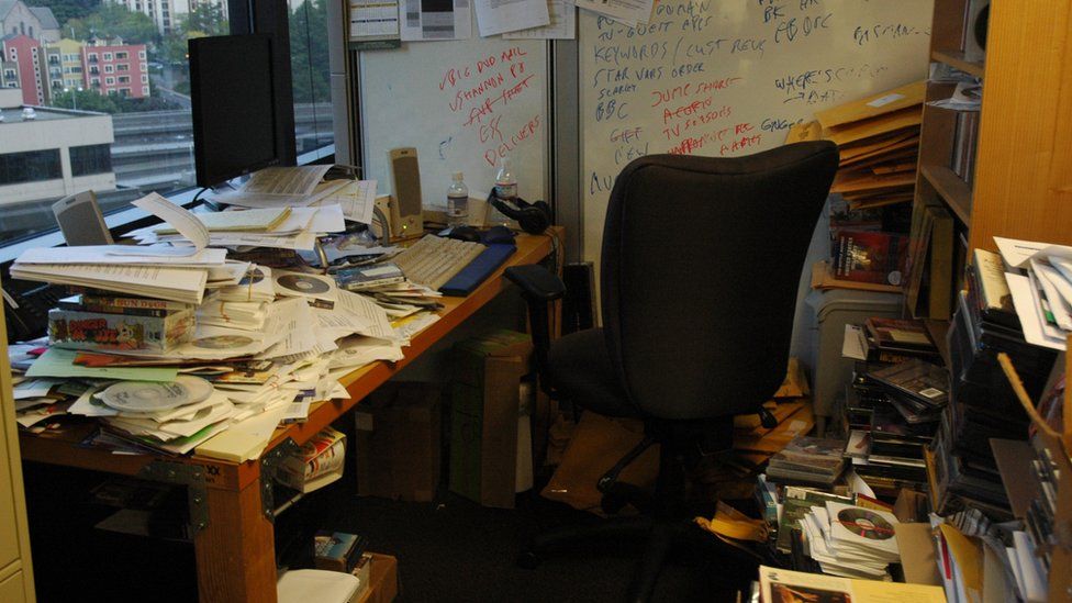 A cluttered home office with piles of newspapers and documents covering the desk and every surface in the small room